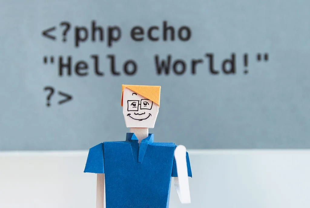 A paper figure in front of a sign reading <?php echo "Hello World!" ?>