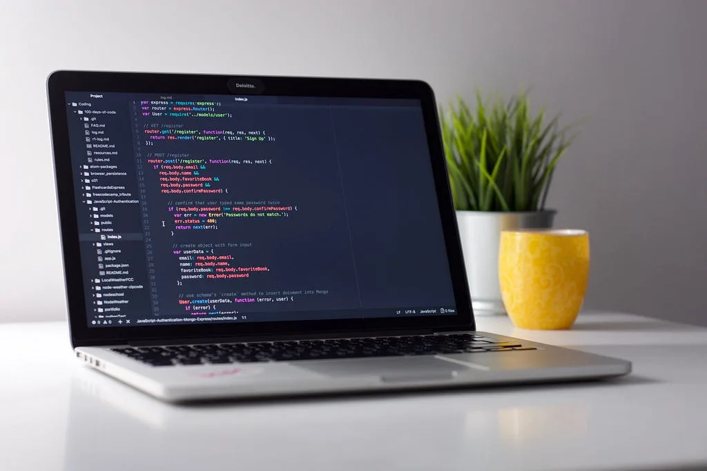 A laptop showing code on a desk beside a plant and a yellow mug